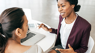 Two women discussing something while sitting next to each other at a circular table in front of a laptop 