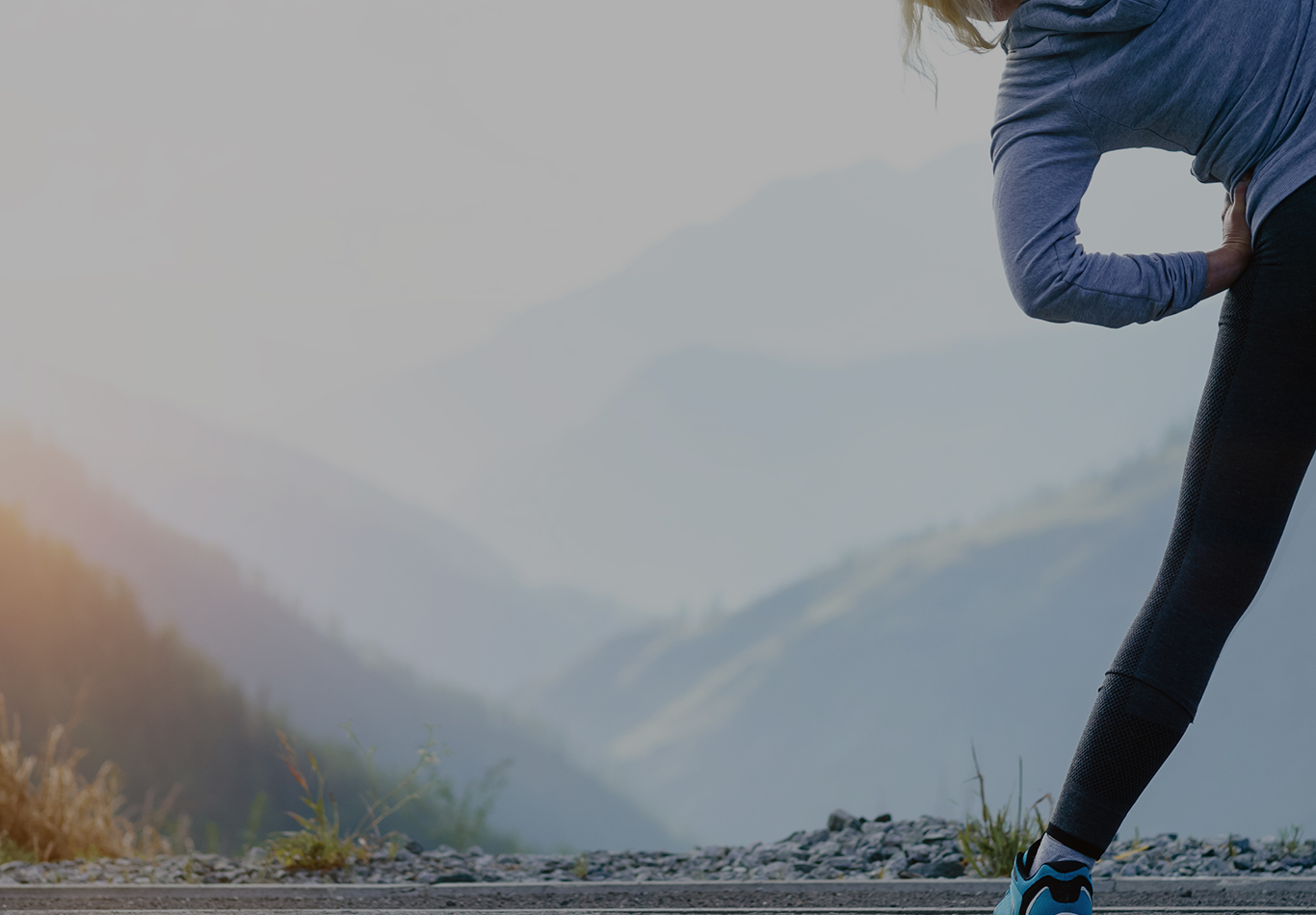 A woman stretching before a run, she is facing towards a scenic view of mountains in the distance. 