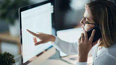 Woman in business attire sitting at a desk, holding a phone, and pointing to a desktop screen.