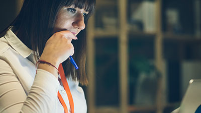 Woman in business attire looking down at laptop screen while holding a pen and hand is touching her face.