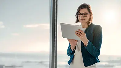 Woman in business attire using tablet while standing next to large windows.