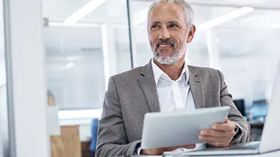 Man in business casual attire sitting while holding a tablet and smiling looking to the left 