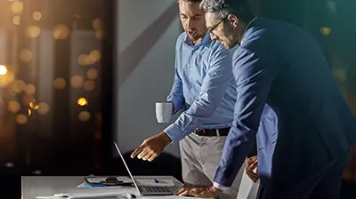 Two men dressed in business casual attire standing while interacting with a laptop.