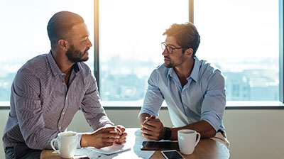 Male colleagues dressed in business casual attire sitting at table and conversing.
