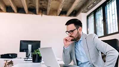A man resting his elbow on a desk and holding his chin while looking at a laptop 