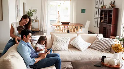 A family sitting on a sectional in their living room. The dad is sitting and holding his daughter while looking at a tablet together. The mother is holding her son while leaning over the couch behind the dad. 