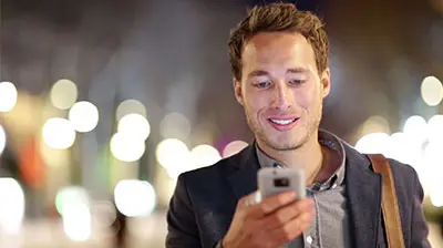 Man in business attire looking down at mobile phone in his hand. There are blurry city lights behind him. 
