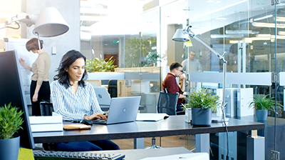Three people in an office setting. Two people are at their desks on their laptops while one person is writing on a whiteboard. 