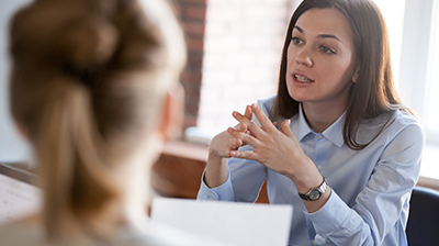 A woman interlocking her fingers in front of her while speaking to other coworkers around a table 