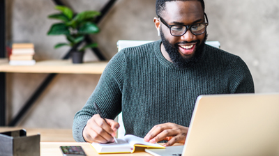 A man smiling and looking at his laptop while writing in a small notebook