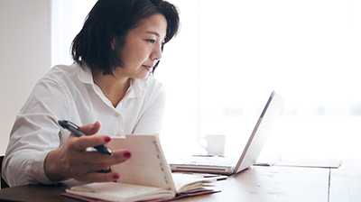A woman sitting at a desk, flipping through her planner while looking at her laptop 