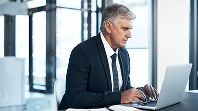 A man wearing a black suit sitting at a desk while typing on a laptop 