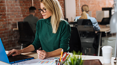 A woman in a green sweater sitting at her desk while typing on a blue laptop 