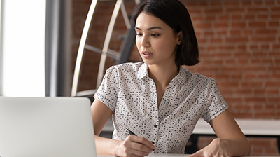 A woman writing something down while looking at her laptop 