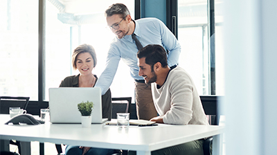Three business people behind a desk. Two of them are sitting down looking at a laptop. One man is standing behind them pointing at the laptop.