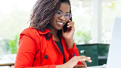 A woman in a red jacket holding a cell phone to her ear while using her laptop
