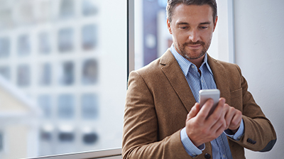 Man in business attire standing in front of a window, looking down at a mobile phone in his hands. 