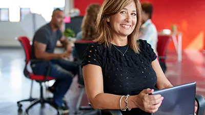 Woman in business casual attire holding a tablet while smiling and looking forward.