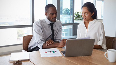 A man and a woman sitting at a table in an office while working on a shared laptop 