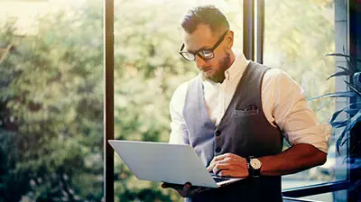 Man in casual attire holding a laptop in both hands and looking down at the laptop screen.
