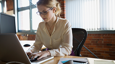 Woman dressed in business attire sitting at desk typing on a laptop.