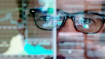 Close up image of man wearing glasses looking straight forward. The reflection of a computer screen can be seen in his glasses.