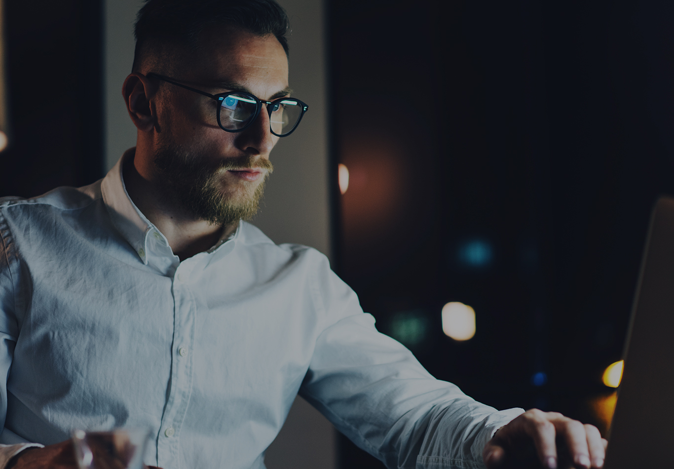 A man with blonde hair, glasses, and facial hair is working on a laptop at night