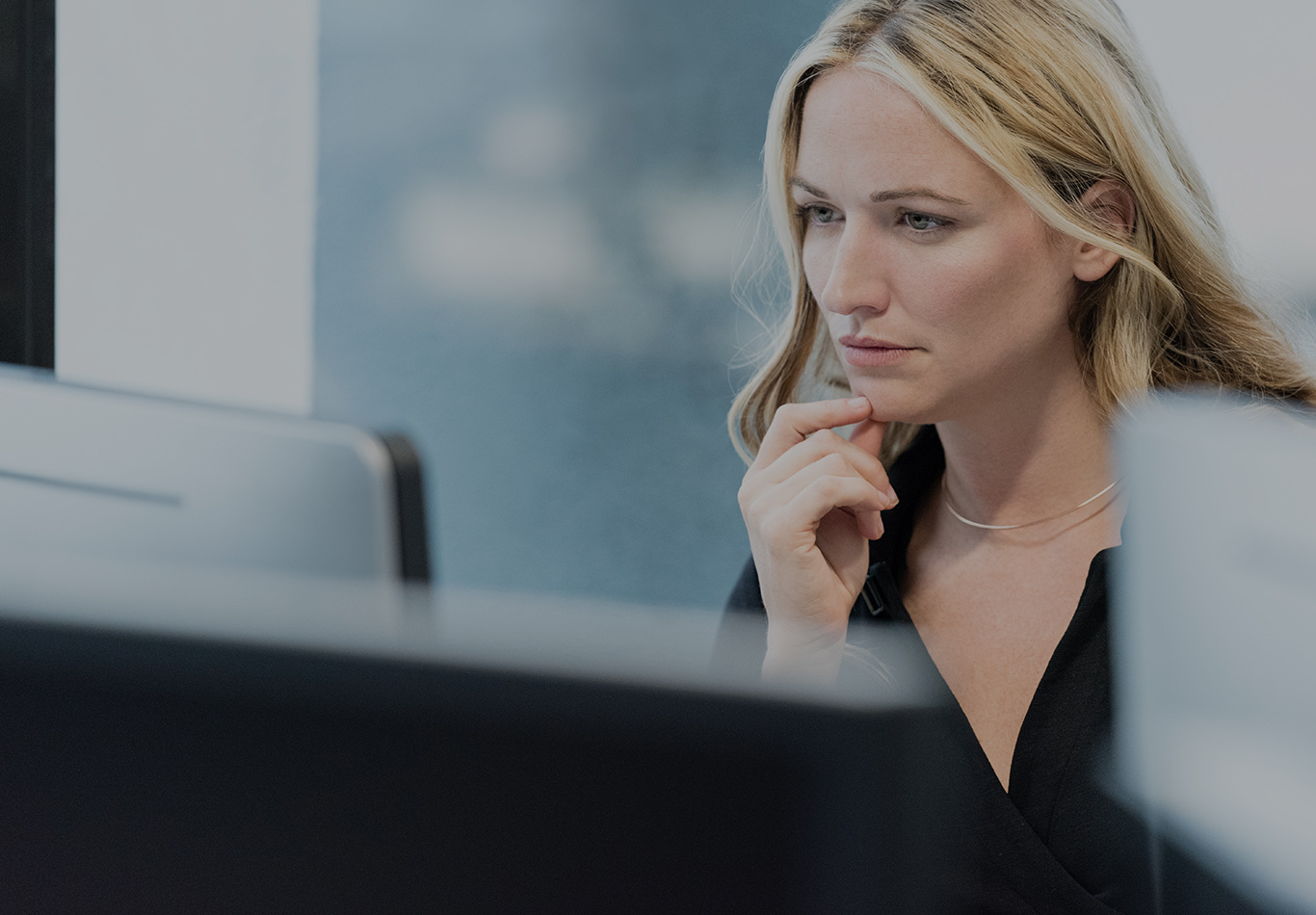 A blonde haired woman with two fingers on her chin is looking at a computer