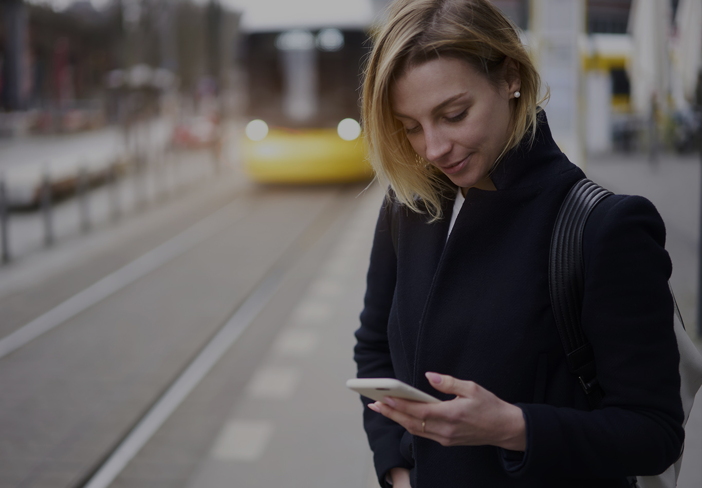 A woman is looking at her phone on the street while a street car is coming down the tracks