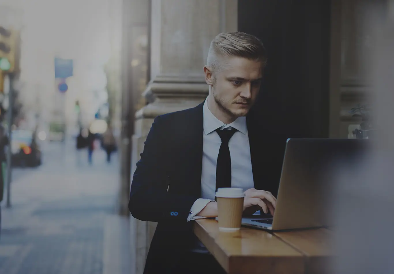 A blonde haired man in business attire is sitting at an outdoor cafe and working on a laptop