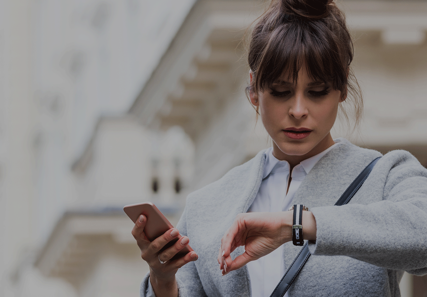 Woman in business attire looking down at watch and holding a smartphone.