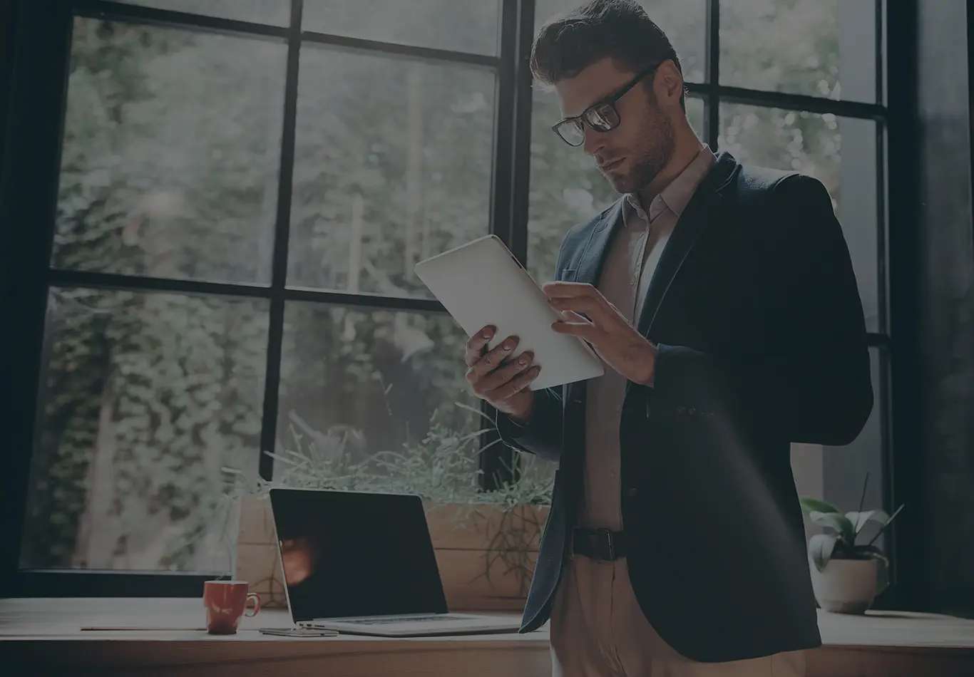 A man with brown hair and glasses is standing up and looking at a tablet. 