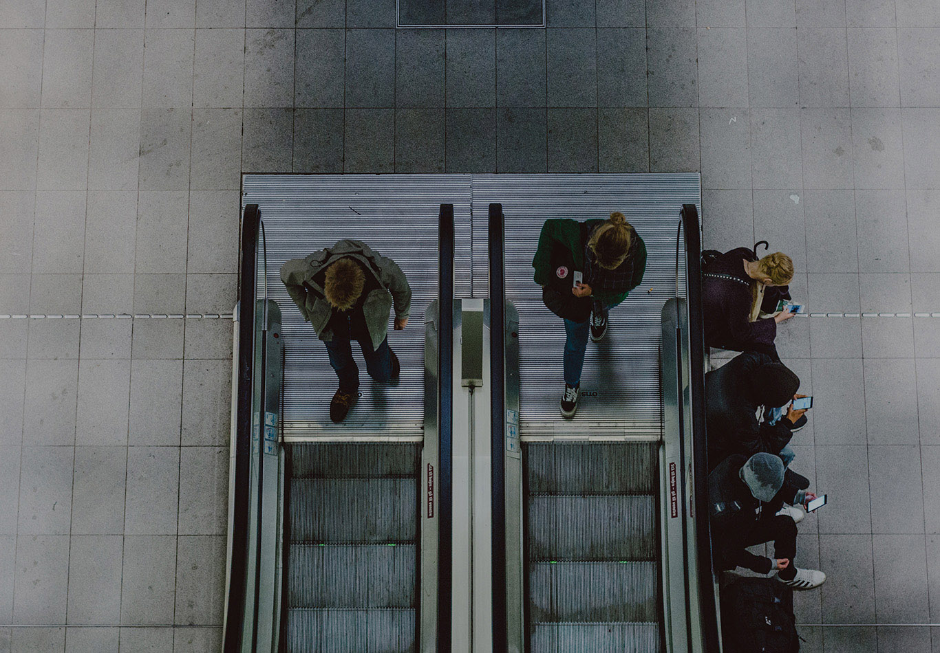 Two people are walking on to two separate escalators. Four people are sitting next to an escalator and they are on their phones