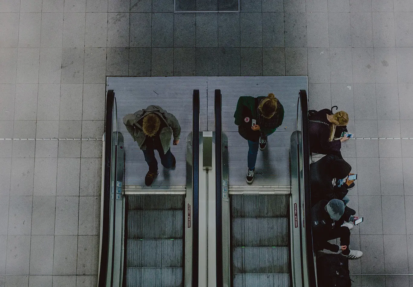 Two people are walking on to two separate escalators. Four people are sitting next to an escalator and they are on their phones
