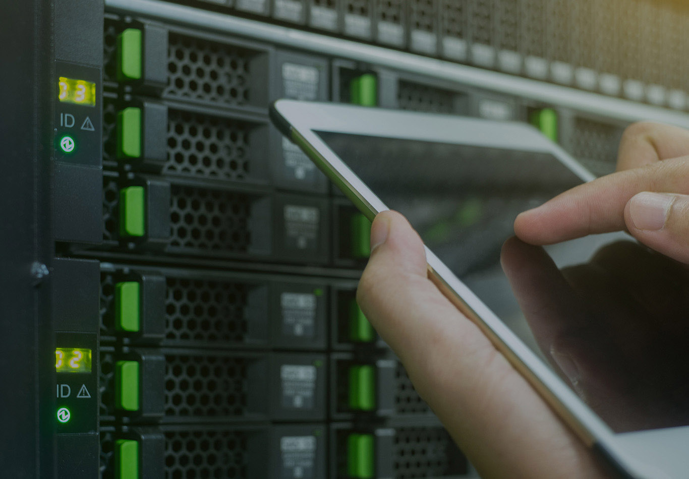 A pair of hands is holding a tablet next to a server rack, 