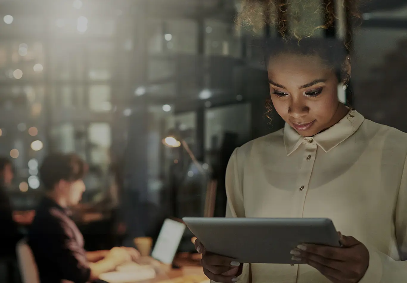 A woman with curly brown hair and a white shirt is looking at her tablet. A second person is out of focus in the background