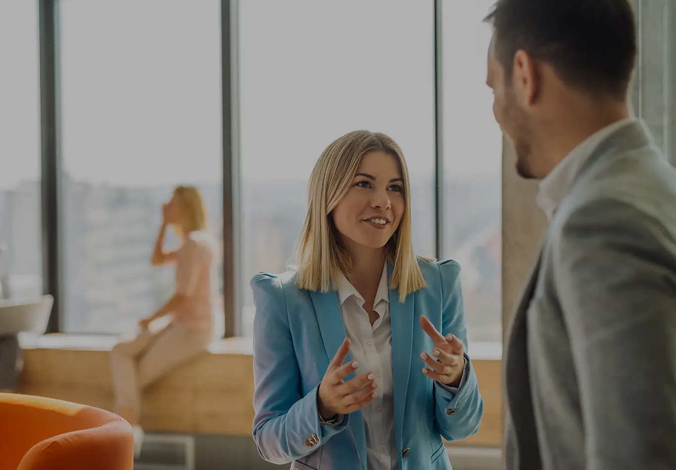 A blonde haired woman and her colleague are discussing something. An out of focus person is sitting by a window