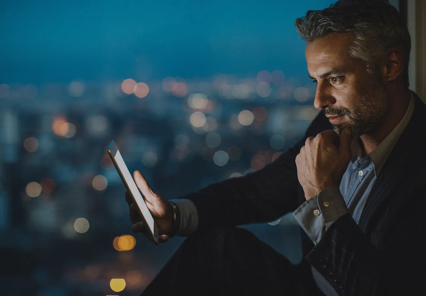 A man is sitting down by a window at night looking at a tablet