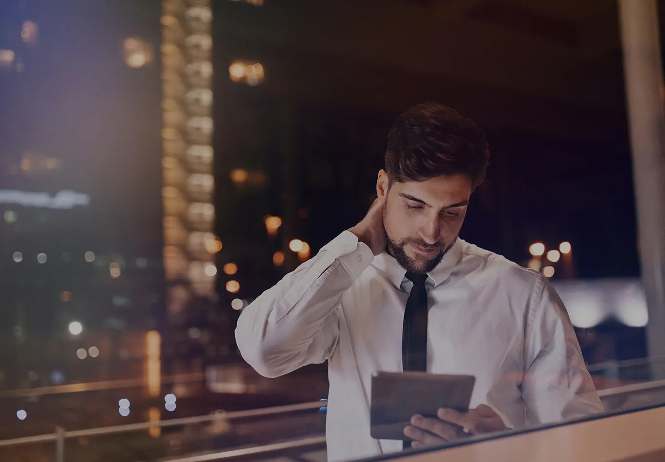 A man is looking at a tablet by a window at night while the street lights are reflected in the window