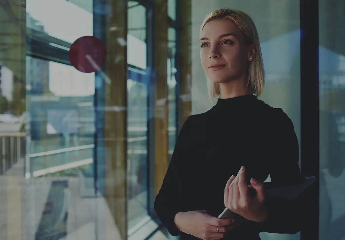 A woman with blonde hair is standing by a window cradling a laptop in her arms