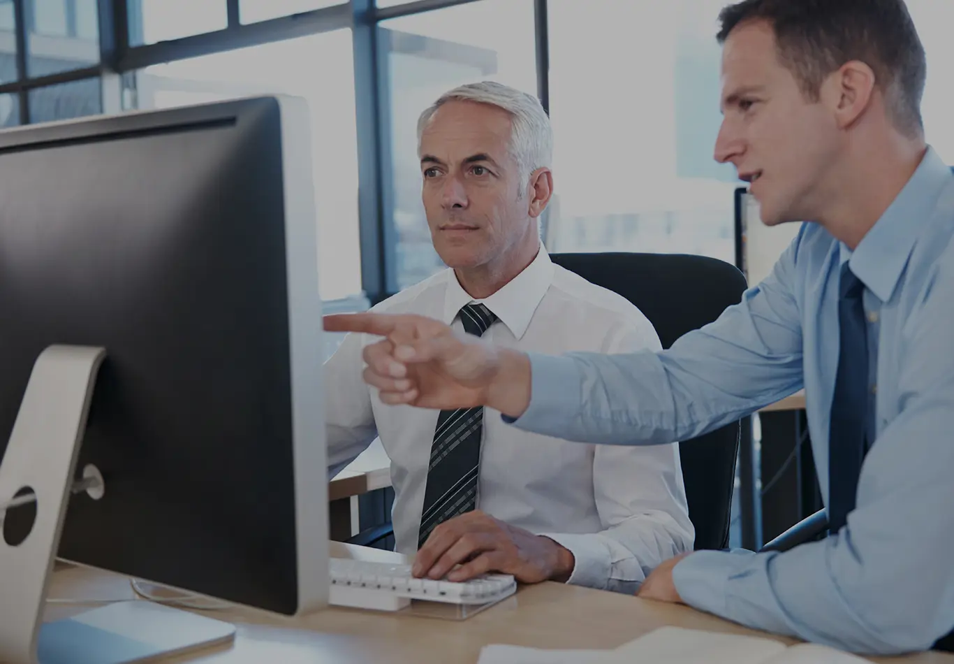 An older man and a younger man are working at a computer together. The younger man is pointing at the monitor