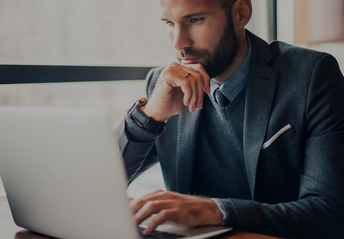 A man with short brown hair and facial hair wearing a suit, sweater and tie is working on a laptop