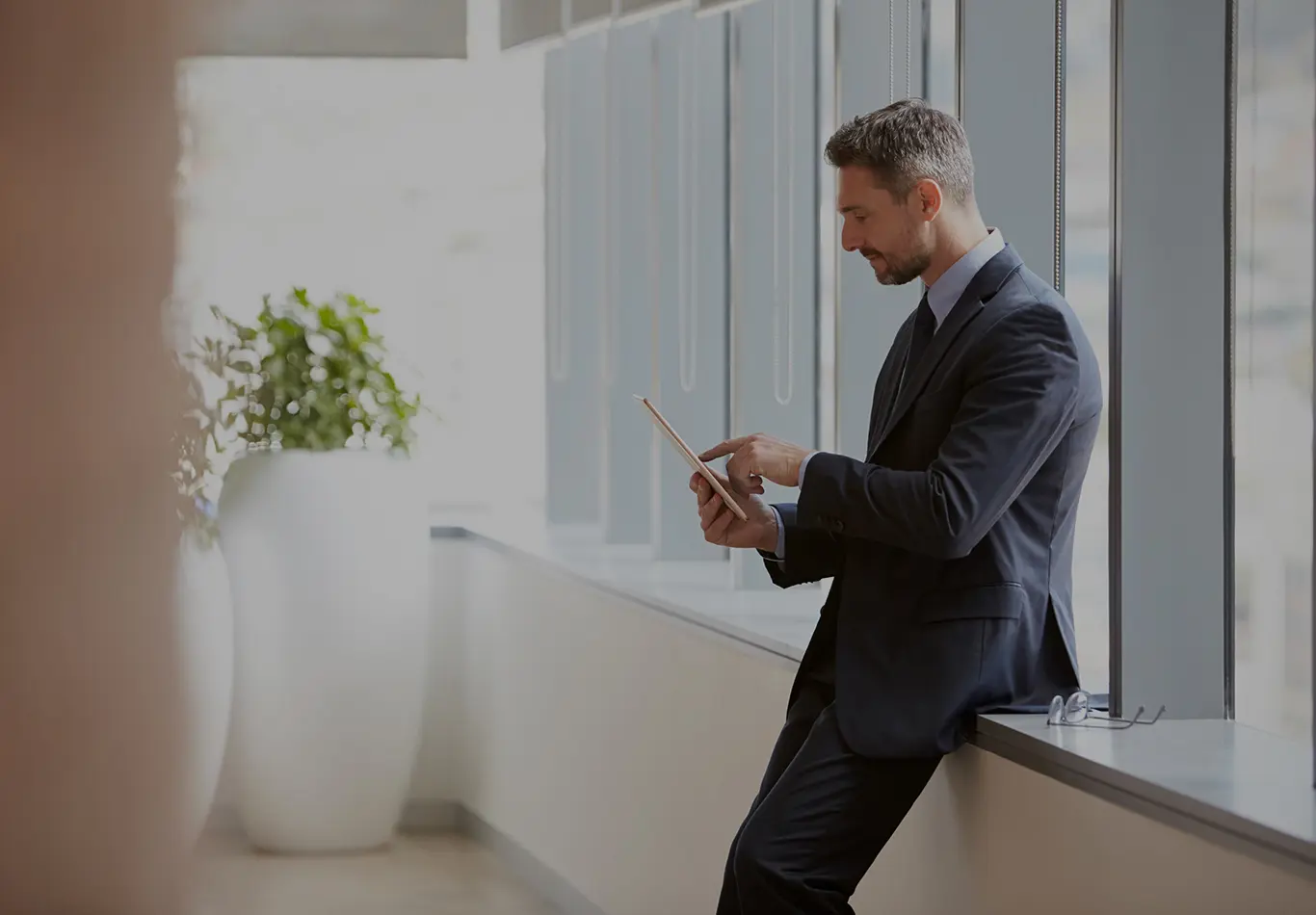A man in a suit is sitting on a window ledge playing with his tablet. 