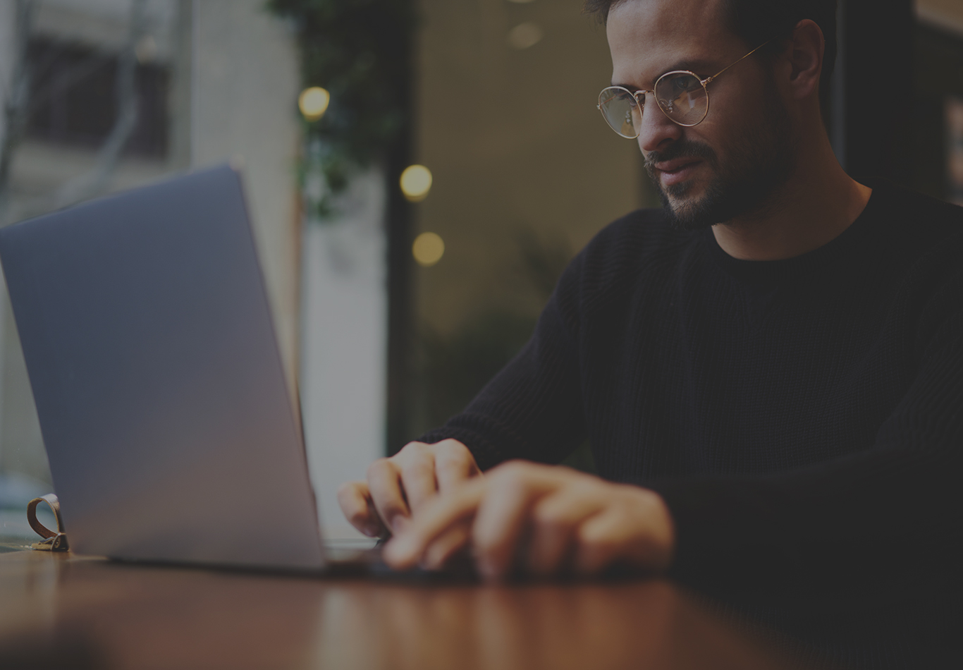 A bearded man wearing circular glasses is working on a laptop