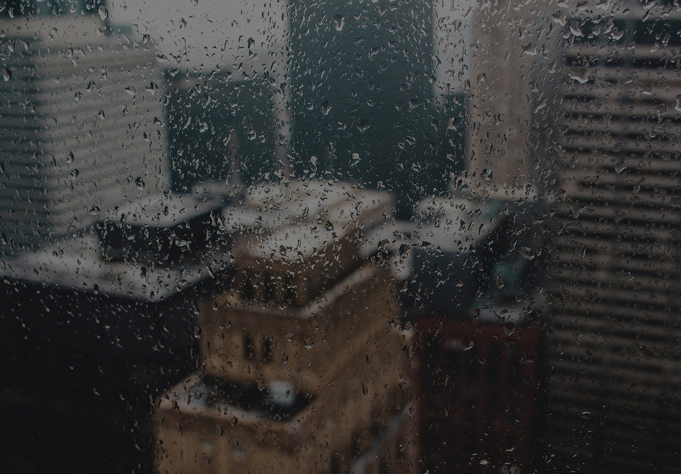A window with some rain drops on it with some buildings out of focus in the background.