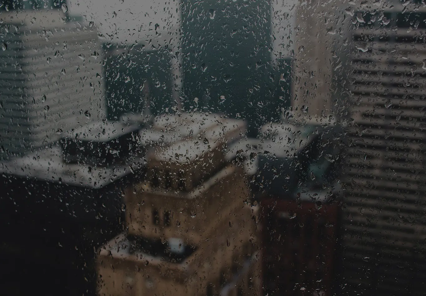 A window with some rain drops on it with some buildings out of focus in the background.