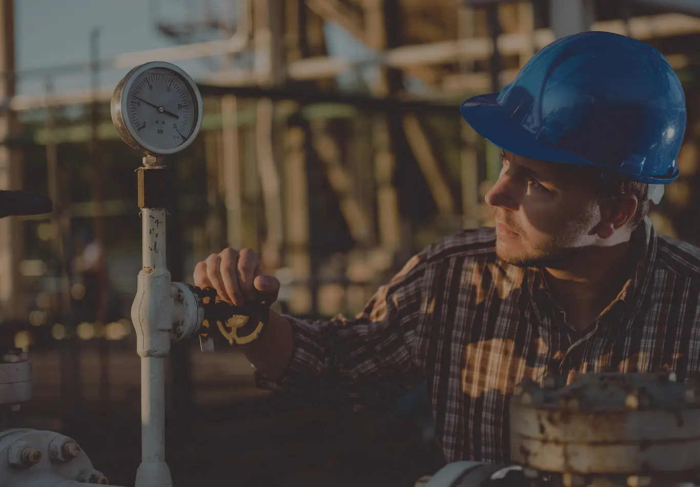 A man with a blue hardhat on is reading a meter on a pipe 