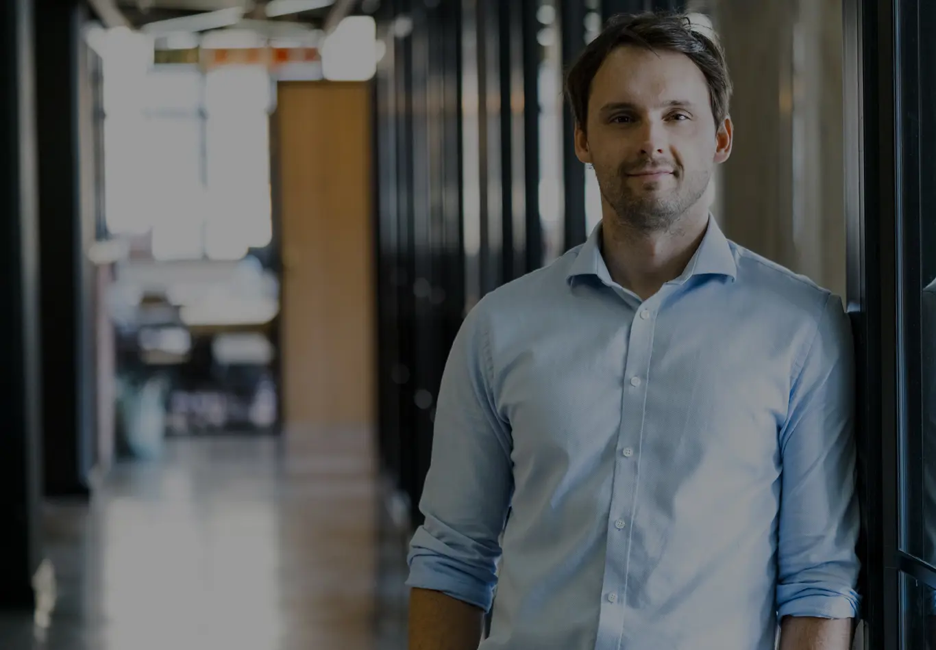 Man in casual attire facing forward and smiling, there is an office in the background. 
